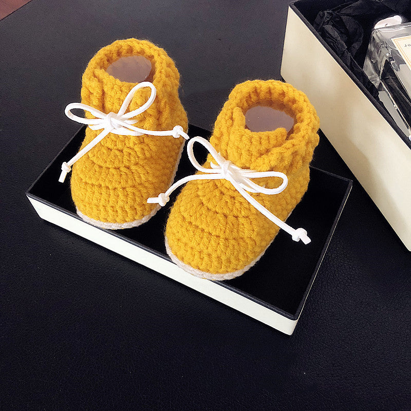 Yellow crocheted baby booties with white laces are neatly placed in an open black and white box, sitting on a black surface.