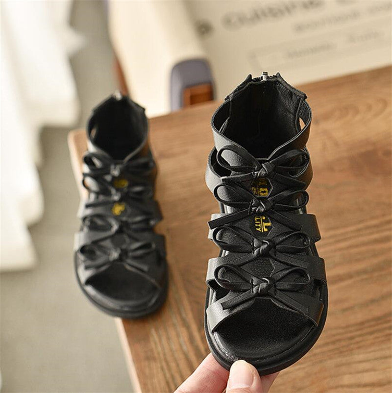 Black leather sandals with bows and a zipper on the back, displayed on a wooden surface. The front is open-toed and features intricate strap detailing.