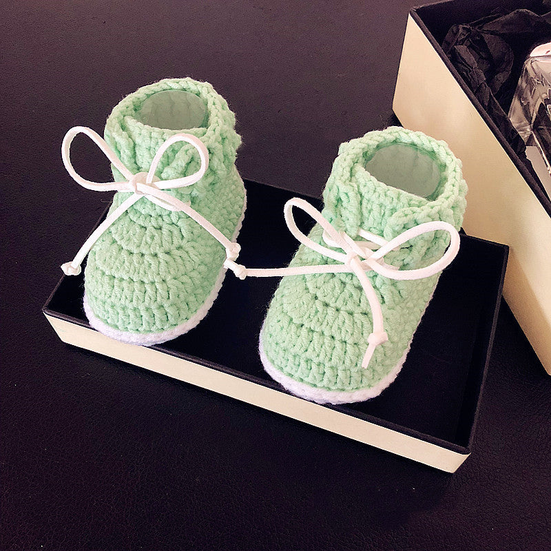 Crocheted baby booties in pastel green with white laces, displayed in a black box on a dark surface. The booties appear soft and cozy, suitable for a newborn.
