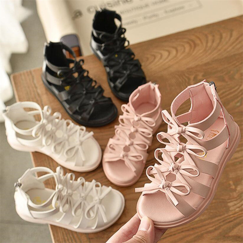 Three pairs of stylish kids' sandals in black, white, and pink with ribbon laces are displayed on a wooden surface. A hand holds the pink pair in the foreground.