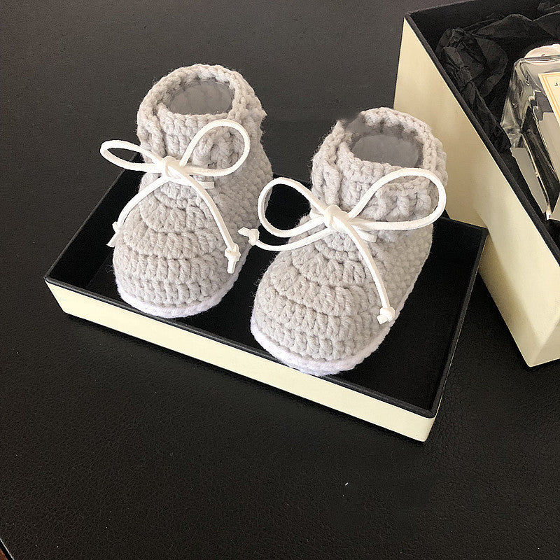 Gray crocheted baby booties with white laces sit on a black gift box. The background includes a partially visible gift box with black tissue paper.