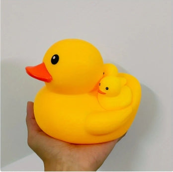 A person holds a bright yellow rubber duck with two smaller ducklings on its back. The large duck features a red beak and round black eyes, and the background is a plain white wall.
