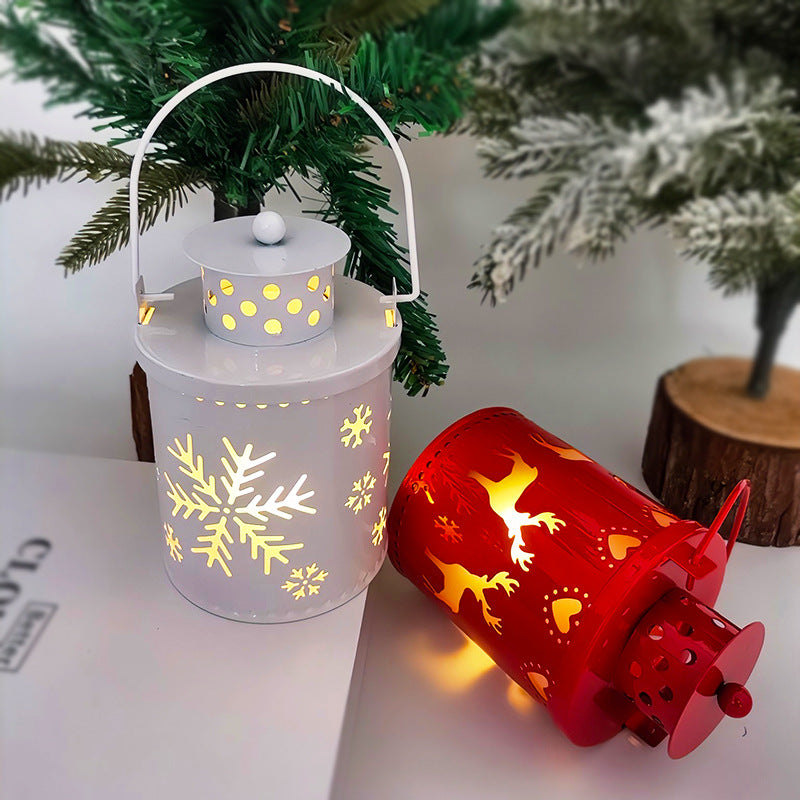 Two festive lanterns on display: a white one with illuminated snowflakes and a red one featuring glowing reindeer silhouettes. They rest under a green pine branch.