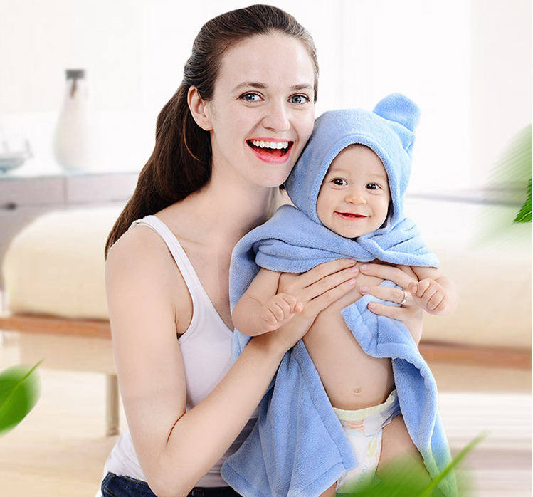 A smiling woman holds a baby wrapped in a blue hooded towel in a bright, cozy room. The baby looks content and is dressed in a diaper. Green leaves blur in the foreground.