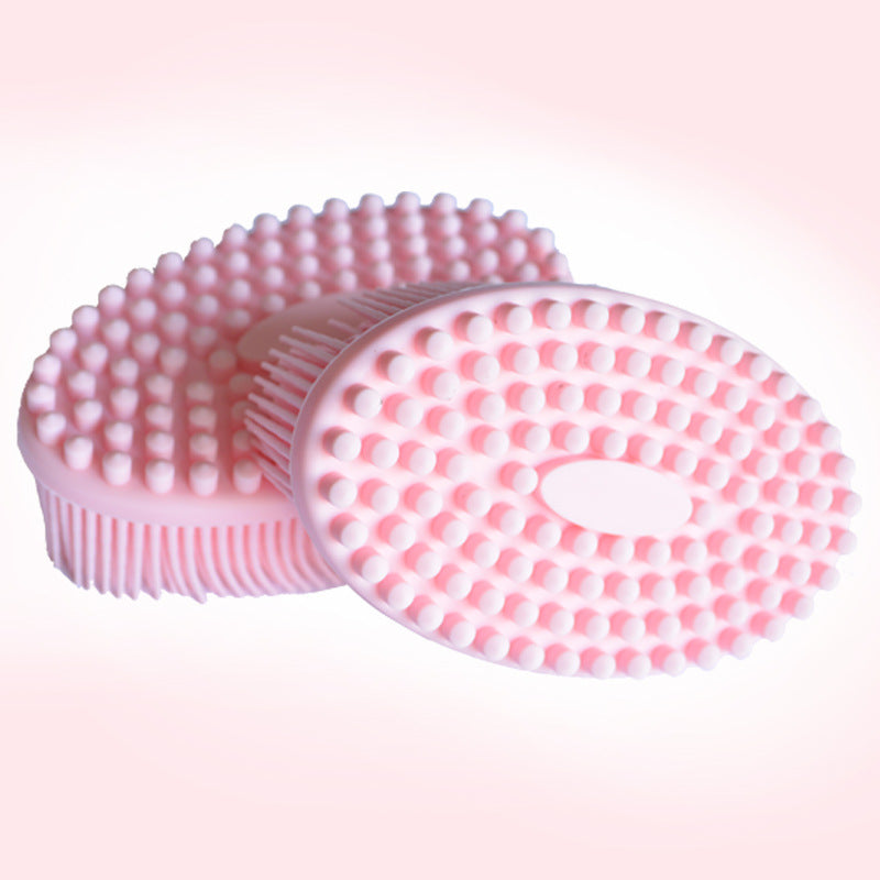 Two pink silicone massage brushes with rounded bristles and oval shapes, placed on a soft pink background. One brush lies flat while the other is tilted, showcasing side bristles.