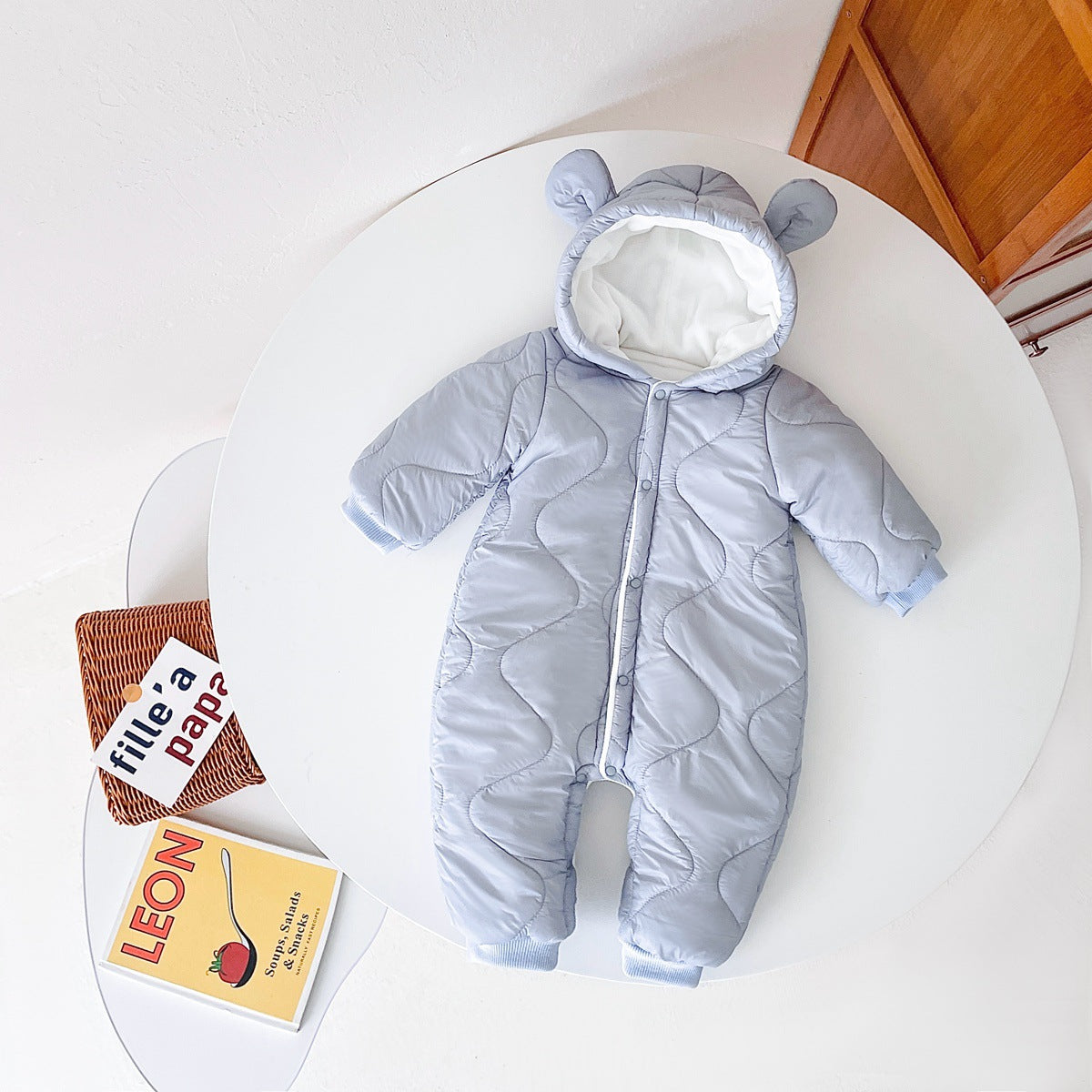 Light blue baby snowsuit with bear ears on the hood, lying on a white table. Books are partially visible at the table's edge.