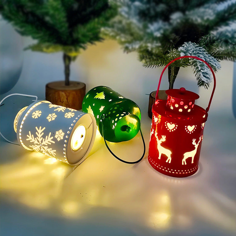 Three decorative lanterns with cutout designs: a white one with snowflakes, a green one with trees, and a red one with reindeer. They're lit and placed near snowy pine trees.