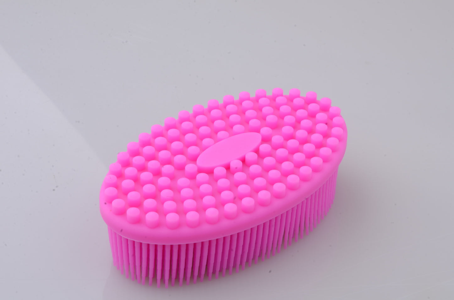 Pink silicone massage brush with an oval shape. It has raised dots on the top side and flexible bristles underneath, laid on a smooth, light gray surface.