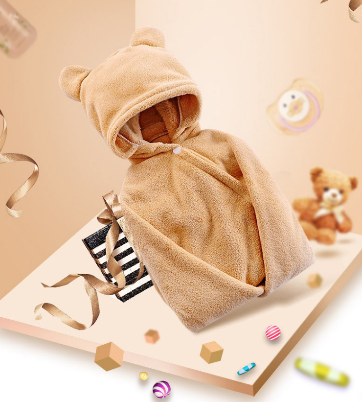 A soft, brown baby hooded blanket with bear ears lies on a striped gift box. Nearby are ribbons, colorful toys, and a blurred teddy bear in the background.