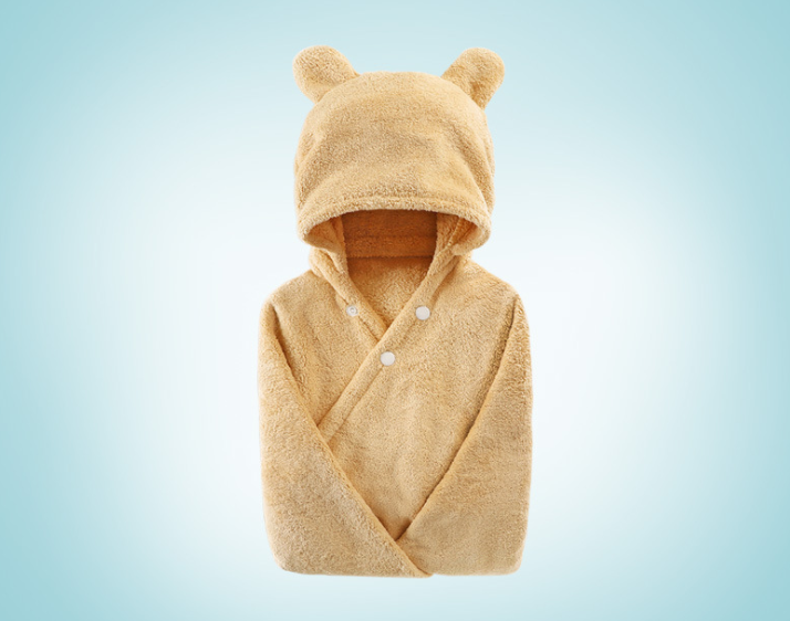 Cozy beige baby wrap with a hood featuring bear ears, buttoned at the front. Made from soft terry fabric against a light blue gradient background.