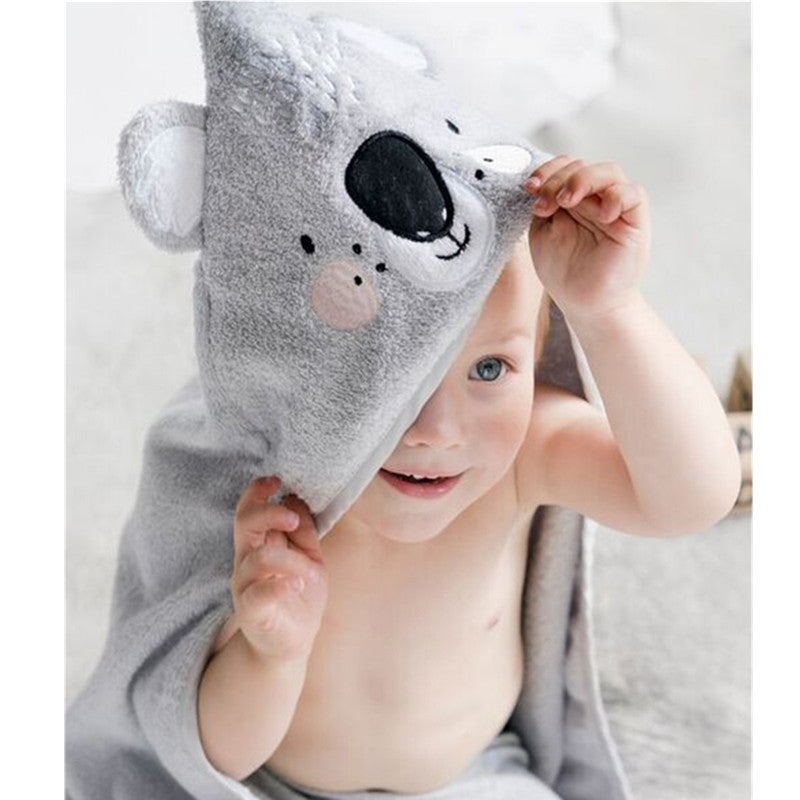 A child peeks from under a cozy gray hooded towel with a cute koala face design, smiling with wide eyes, sitting on a soft surface.