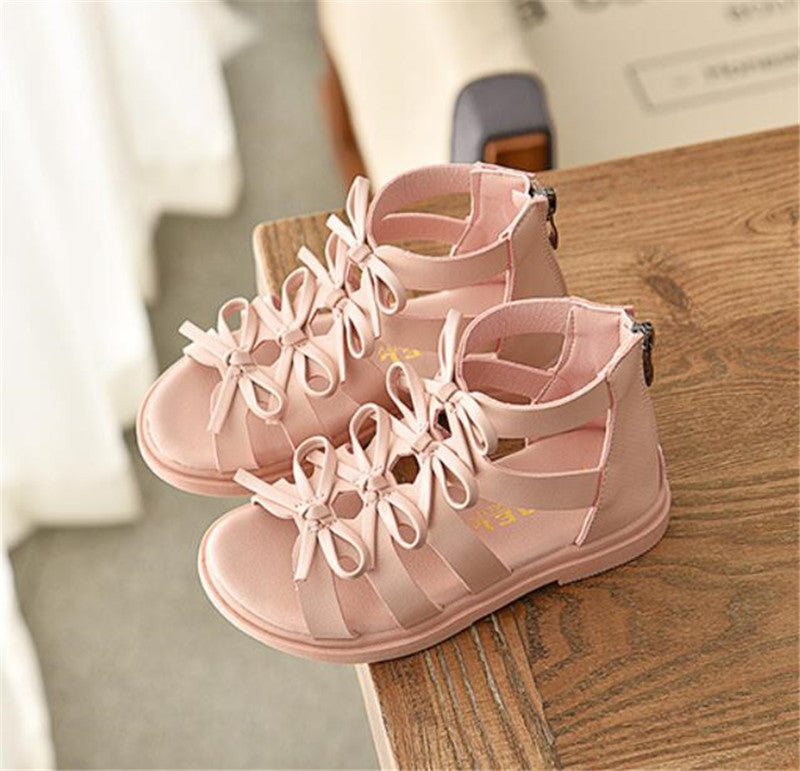 Light pink, open-toe sandals with multiple bow accents, featuring a back zipper. The sandals rest on a wooden surface, with soft, natural lighting.