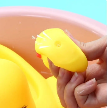 A close-up of a person squeezing a yellow rubber duck with small water jet holes. The duck is held over a pink basin with a turquoise background.