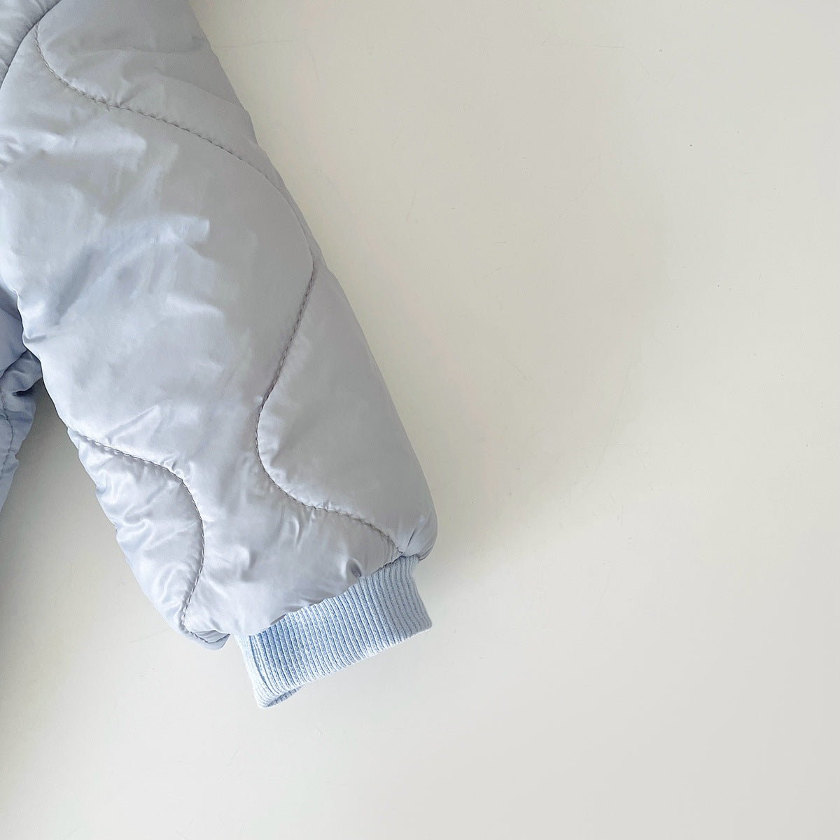 Close-up of a light gray quilted jacket sleeve with ribbed blue cuff, against a plain white background. The stitching forms a curved pattern on the fabric.