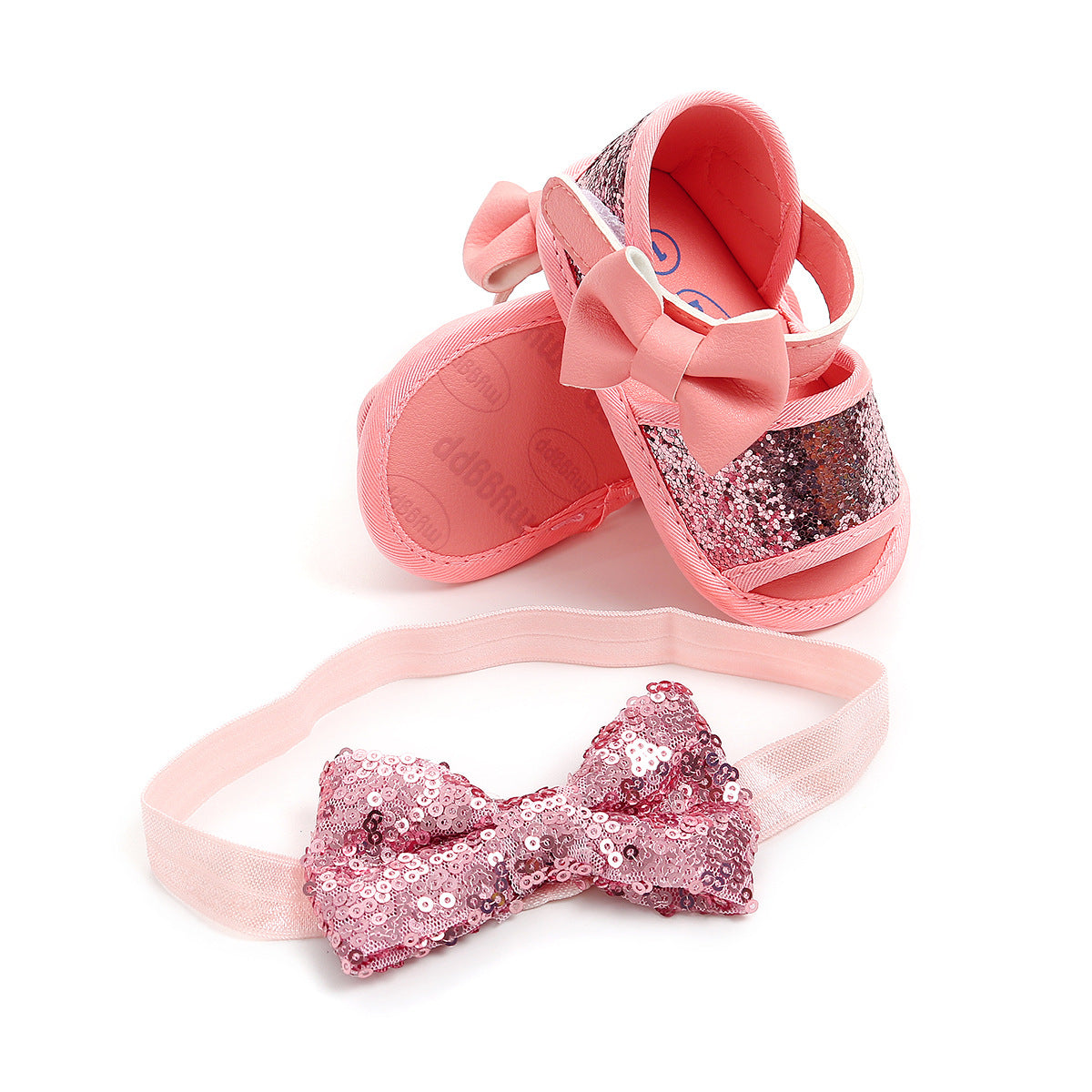 Pink baby shoes adorned with sequins and bows, paired with a matching pink sequined headband, set against a white background.
