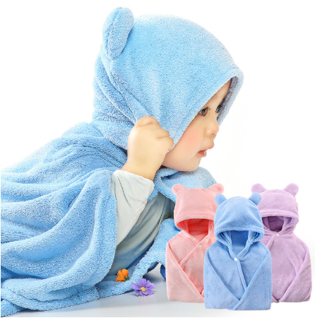 A baby in a blue, bear-eared hooded towel lies beside folded towels in pink, light blue, and purple. Flowers are scattered nearby.