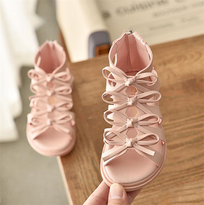 Pink children's sandals with multiple bow straps and open toes, placed on a wooden surface. One sandal is held up for display, showcasing its intricate design.