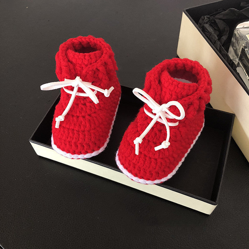 Red crocheted baby booties with white laces on a black surface, partially placed inside an open box. The booties have a white trim along the soles.