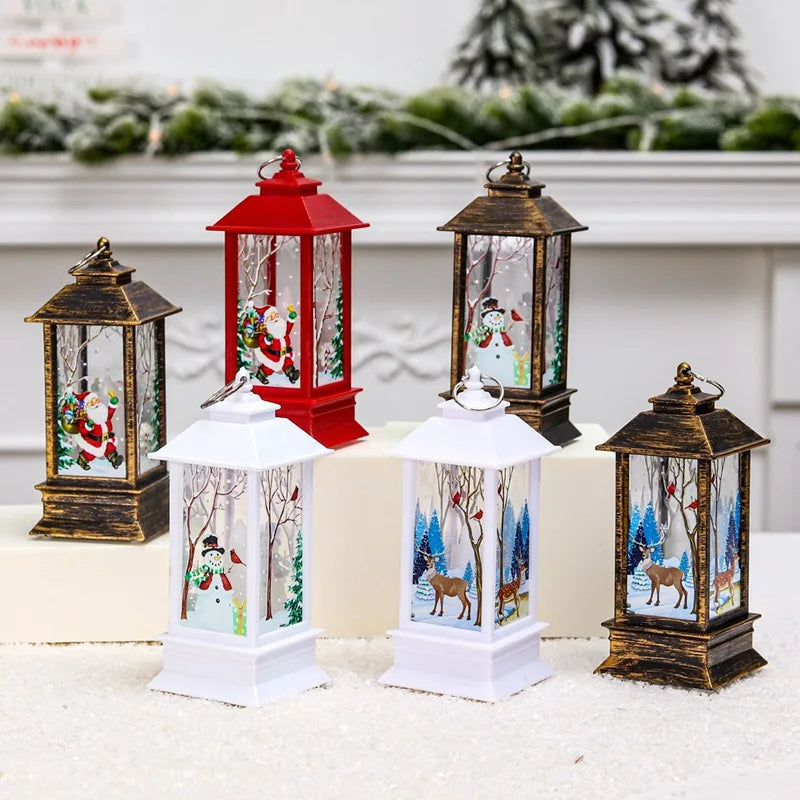 Six festive lanterns with winter scenes, featuring snowmen, Santa, and deer. They are in various colors—white, red, and bronze—set on a snowy surface with green foliage in the background.