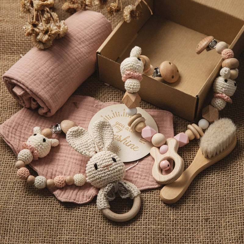 Baby gift set on burlap includes a crocheted bunny teether, pink and beige beads, wooden brush, and a boxed card reading "you are my sunshine." Dried flowers decorate the scene.
