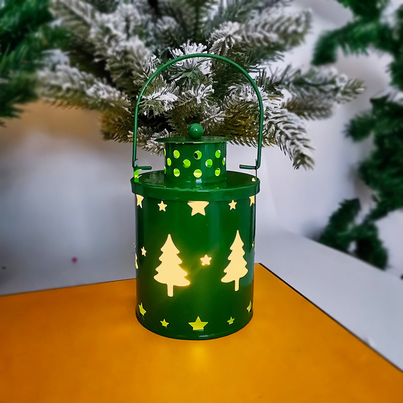 Green lantern with star and tree cutouts, glowing warmly. It sits on a yellow surface, with a snow-frosted Christmas tree in the background.