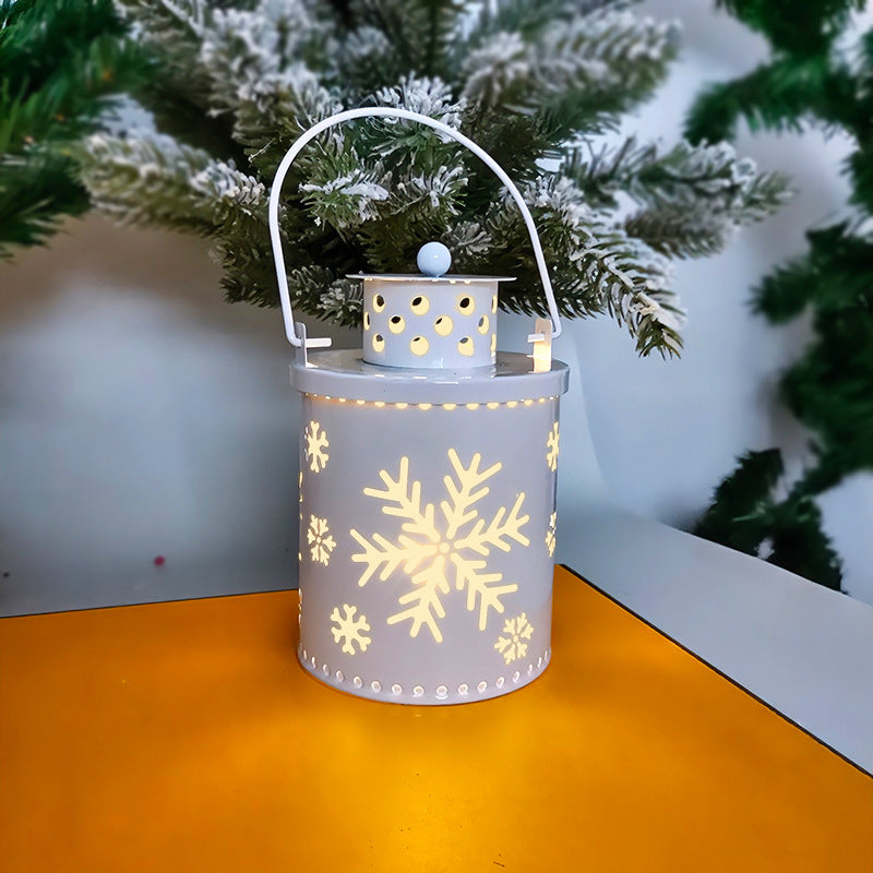White lantern with glowing snowflake cutouts sits on an orange surface, with a snowy evergreen tree in the background, creating a cozy winter ambiance.