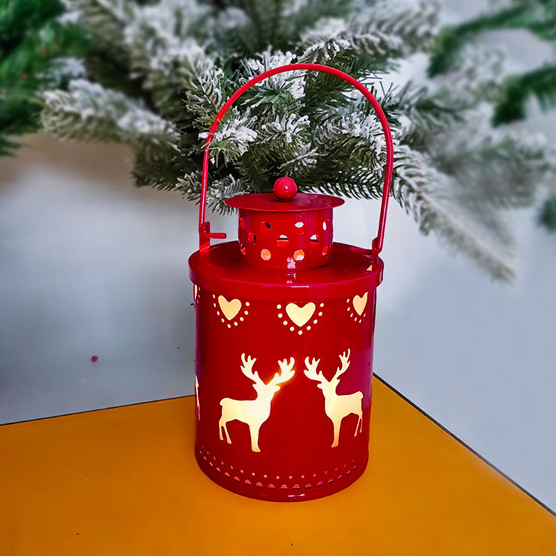 A red lantern with reindeer and heart cutouts is lit, casting warm light. It sits on a yellow surface with snowy pine branches in the background, creating a festive scene.