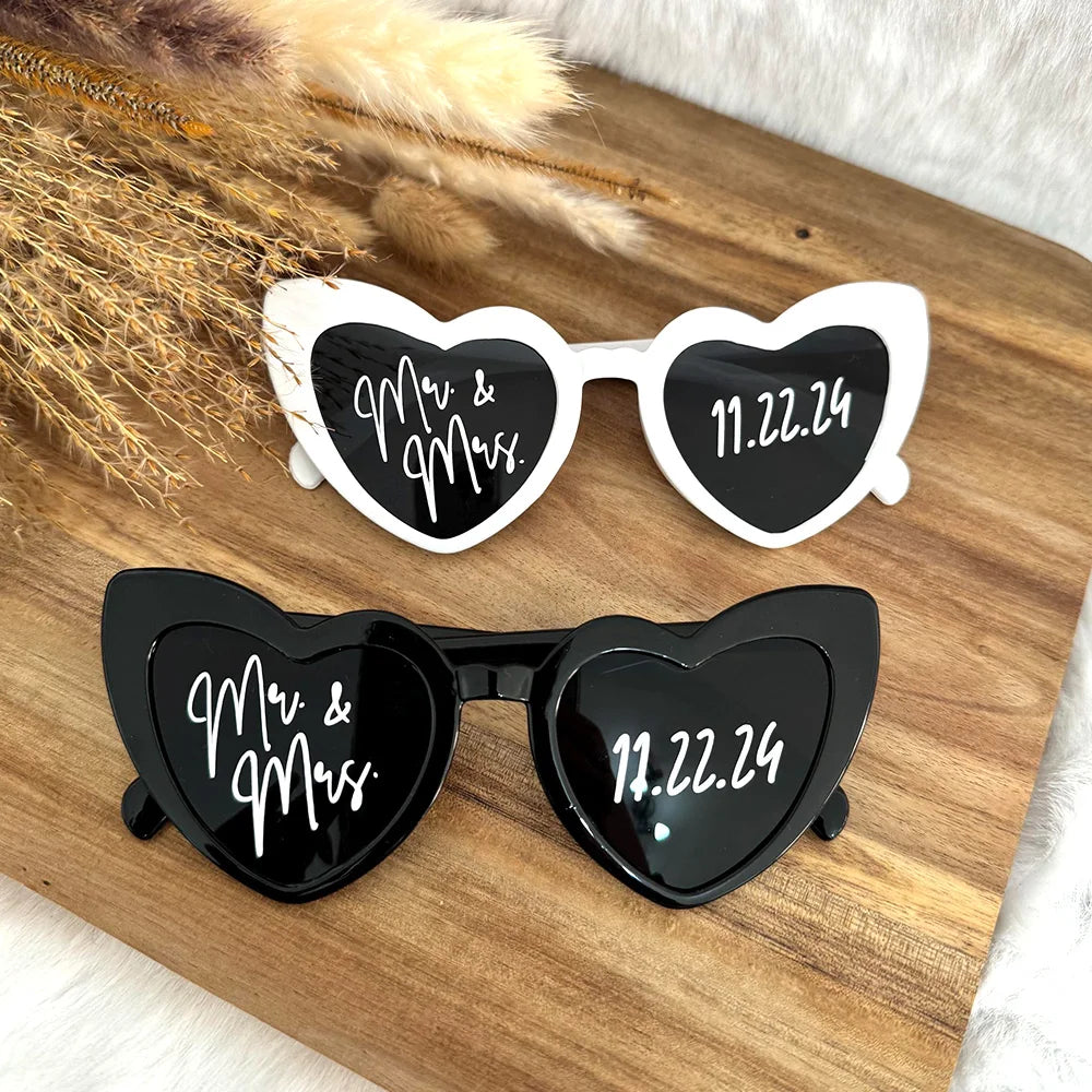Heart-shaped sunglasses with "Mr. & Mrs." and "11.22.24" on the lenses, one pair in black, one in white, resting on a wooden surface with dried plants.