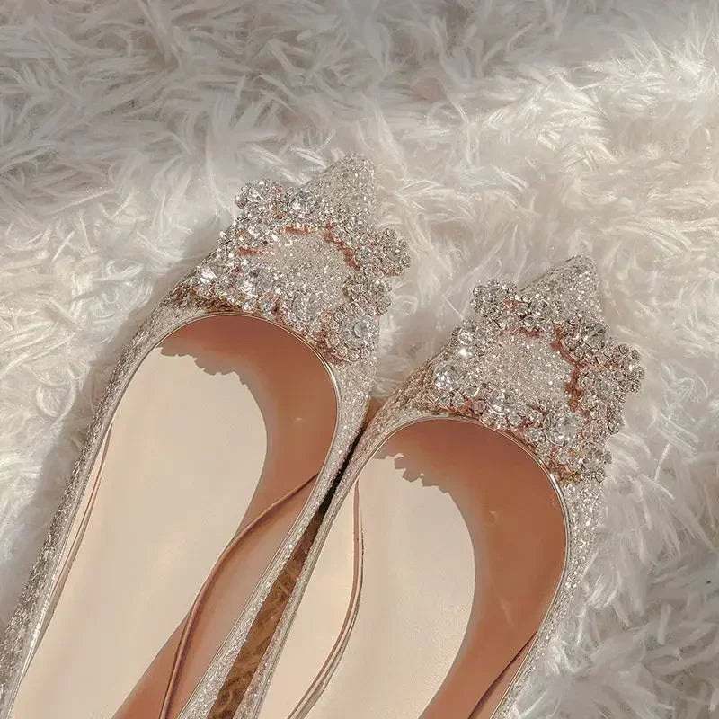 Sparkling pointed-toe women's shoes adorned with intricate rhinestones, lying on a fluffy white surface. The shoes have a glossy, elegant finish with a nude interior.