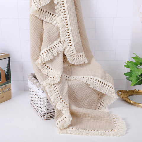 A soft beige blanket with crochet edges draped over a white wicker basket. Nearby, a green plant and a book provide a cozy, elegant setting on a light-colored surface.