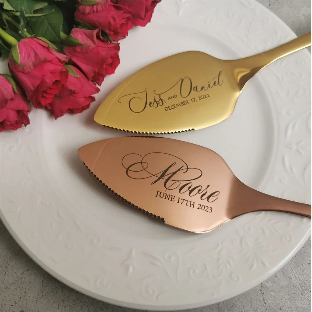 Gold and copper cake servers engraved with names and dates rest on a white plate. A bouquet of red roses decorates the top left corner, adding a romantic touch.
