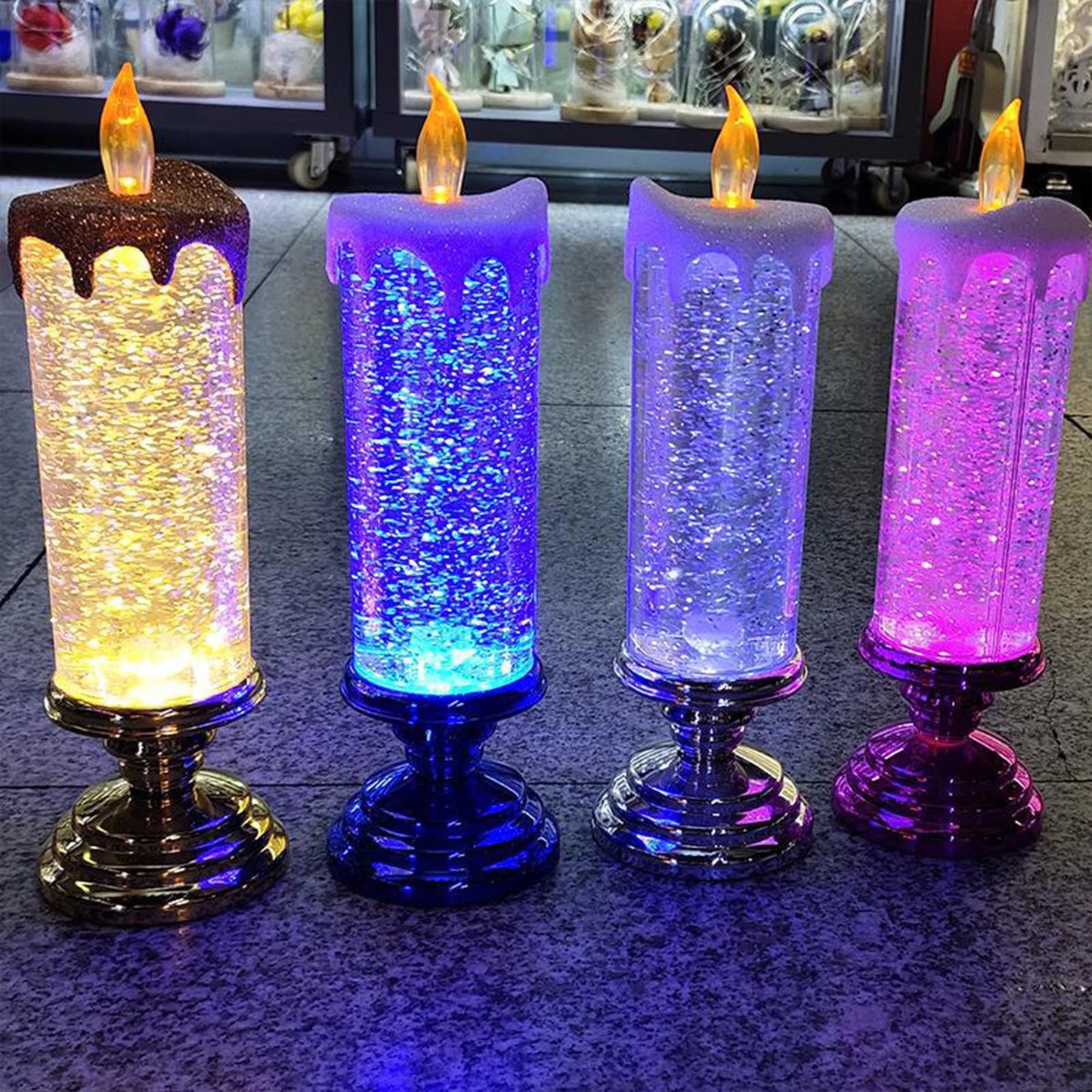 Four LED candles on silver bases, each with color-changing glitter effects in shades of yellow, blue, white, and pink, standing on a tiled surface against a blurred background.