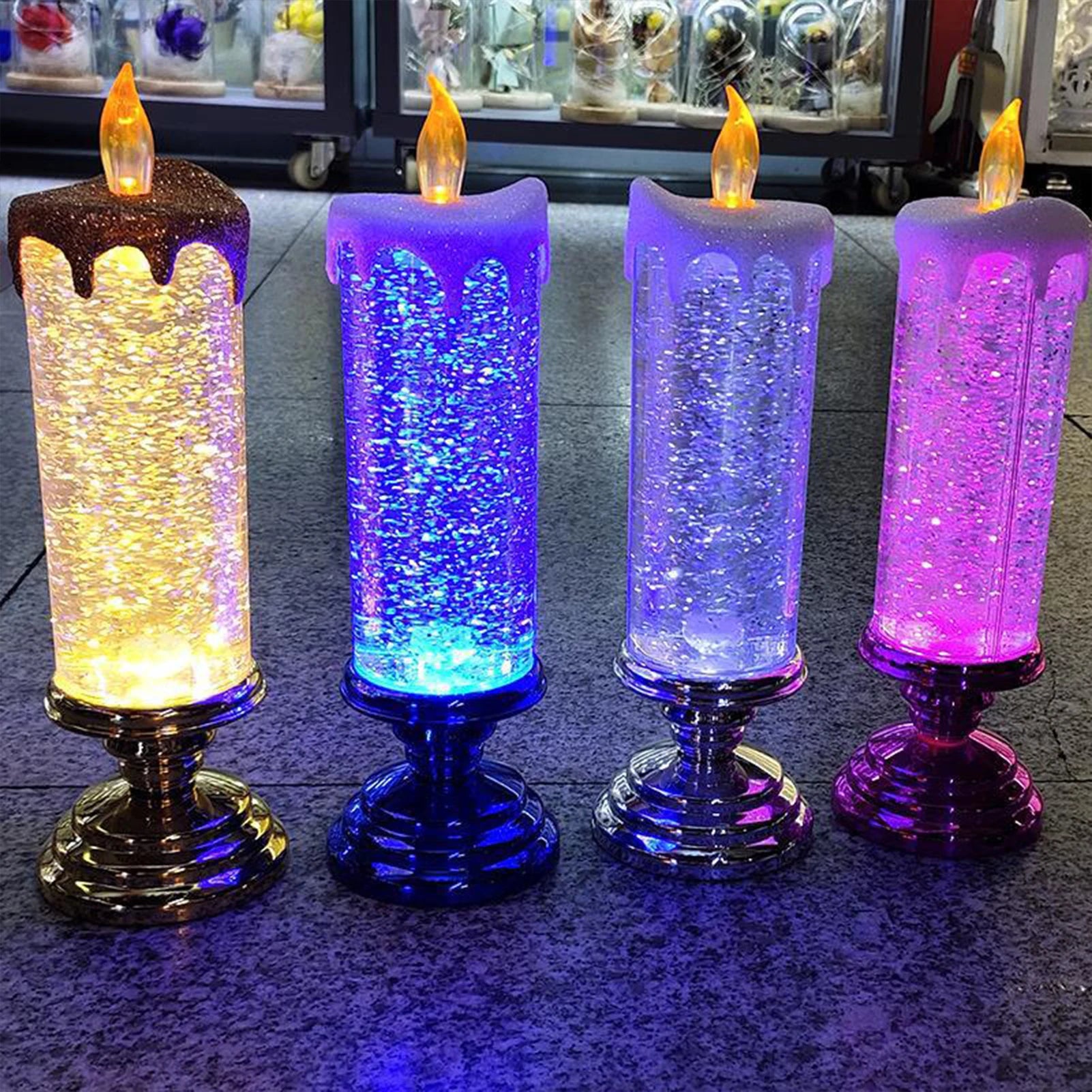 Four LED candles on silver bases, each with color-changing glitter effects in shades of yellow, blue, white, and pink, standing on a tiled surface against a blurred background.