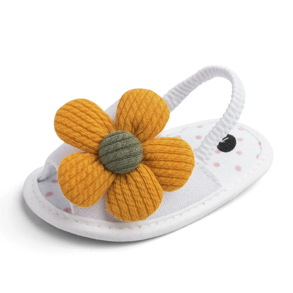White baby sandal with a large yellow fabric flower featuring a green center. It has an elastic strap and a white sole with pink polka dots and black embroidery accents.