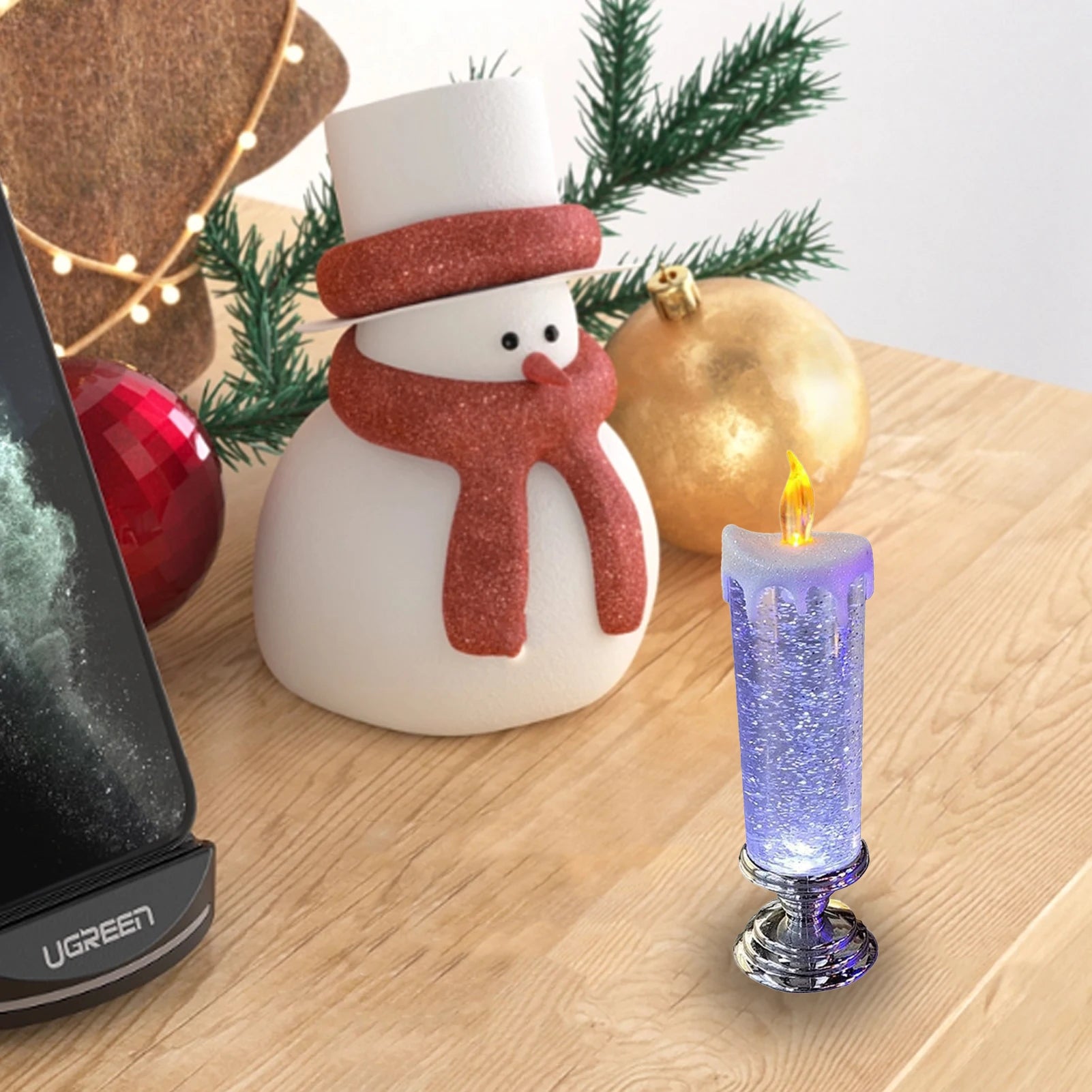 A snowman ornament with a red scarf and hat sits on a wooden table next to a purple glitter candle and Christmas ornaments. A phone on a stand is partially visible.