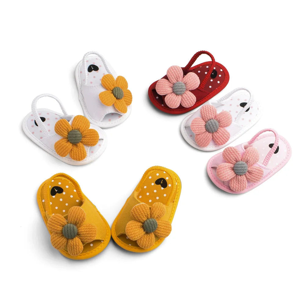 Colorful baby sandals with large fabric flowers: two pairs in yellow, two in white, one in red, and one in pink, arranged in a circle on a white surface.