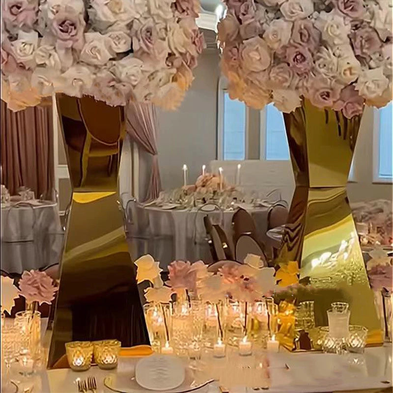 Elegant dining table setting with large golden vases topped with pink and white roses. Candles and glasses create a warm ambiance. Luxurious drapes and chairs in the background.