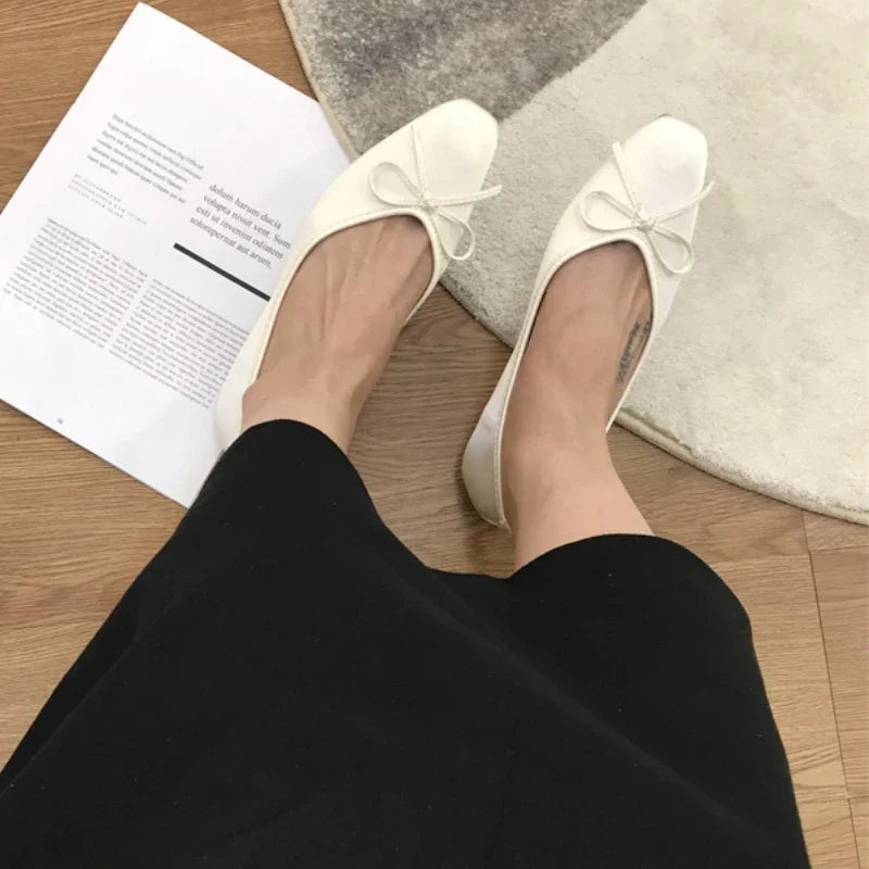 Person wearing white ballet flats with bows, sitting on a wooden floor next to an open magazine. A black skirt is visible, and part of a round rug is on the right.