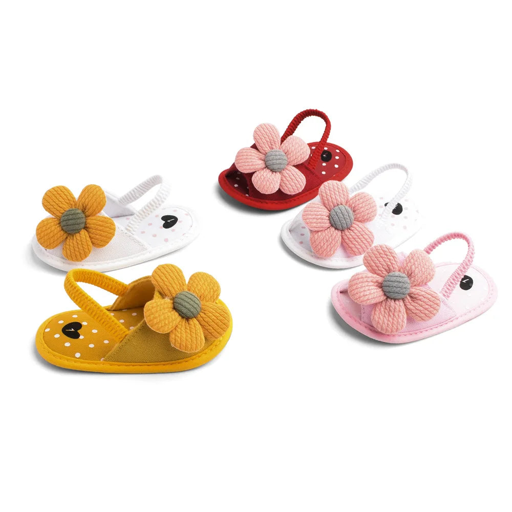Five pairs of baby sandals with thick flower embellishments in yellow, red, pink, and white, arranged in a semicircle on a white background.