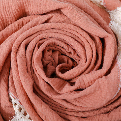 A soft, coral-colored fabric intricately rolled to form a spiral pattern, with fringed edges visible at the top right corner, creating a warm and cozy appearance.