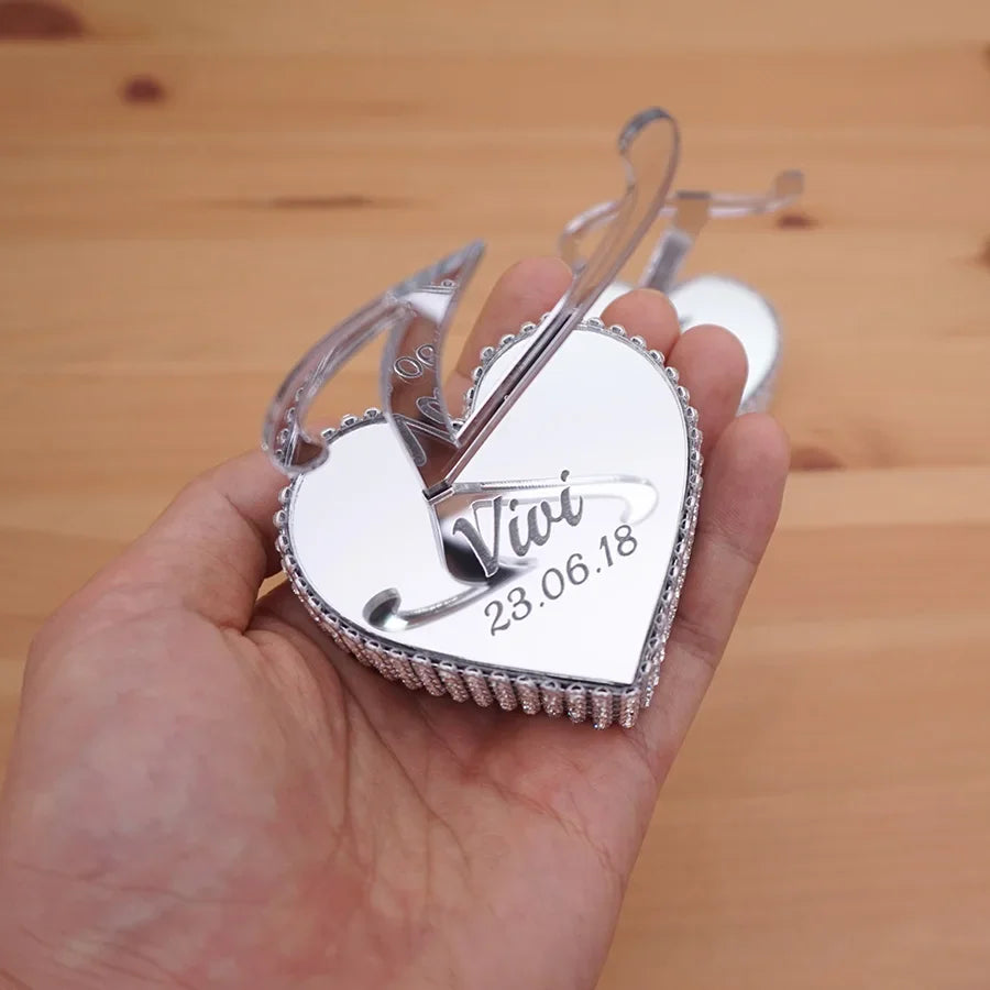 A hand holds a heart-shaped, silver engraved lock with the name "Vivi" and date "23.06.18" on a wooden surface. The lock features a decorative bow design.