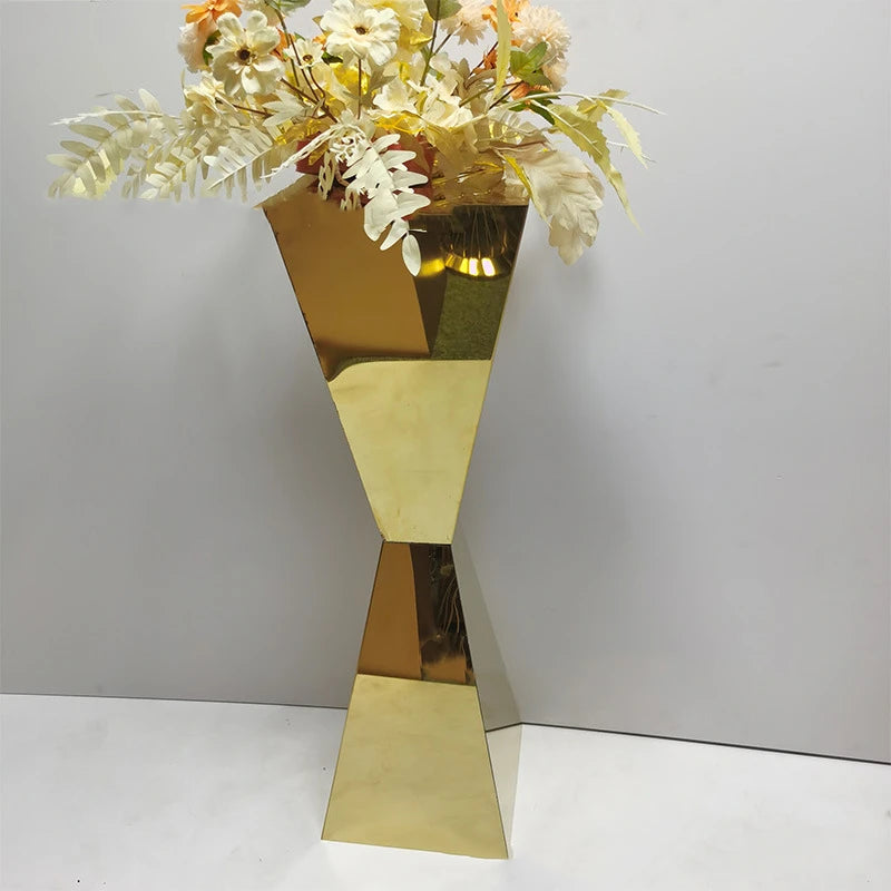Tall, geometric gold vase with an hourglass shape, filled with cream and pale orange artificial flowers and ferns, set against a plain white background.