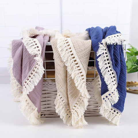 Three woven blankets in purple, beige, and blue with fringed edges draped over a white wicker basket. They are set against a light, tiled background with a small green plant.