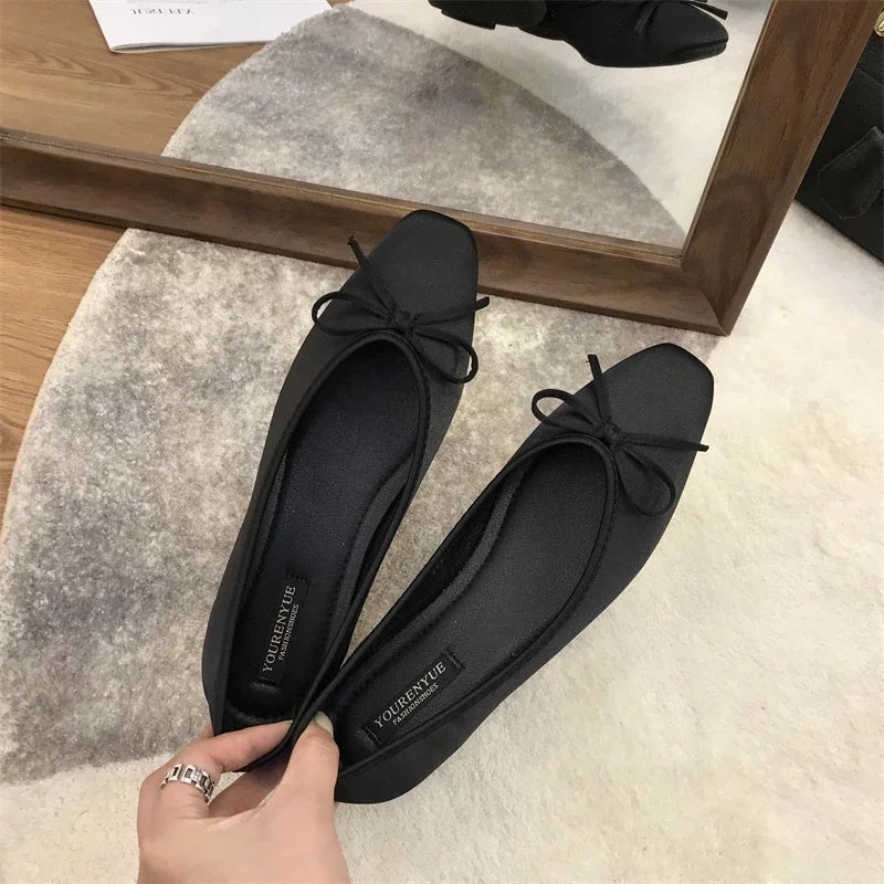 Black ballet flats with bows, held in hand, are reflected in a mirror. They rest on a gray and white rug beside a wooden floor, with a part of a black bag visible.