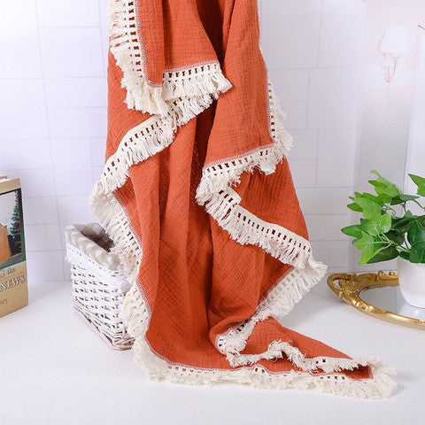 A woven, rust-colored throw blanket with white fringe edges draped artistically over a white basket on a tabletop, accompanied by a small green plant and a book titled "News."