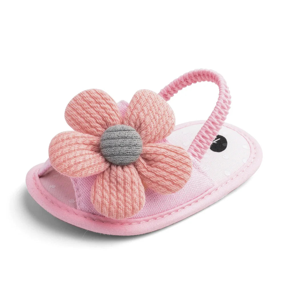 Pink baby sandal with a soft fabric flower on top. The sandal features a textured pink strap and a small eye design near the toe area on a white base.