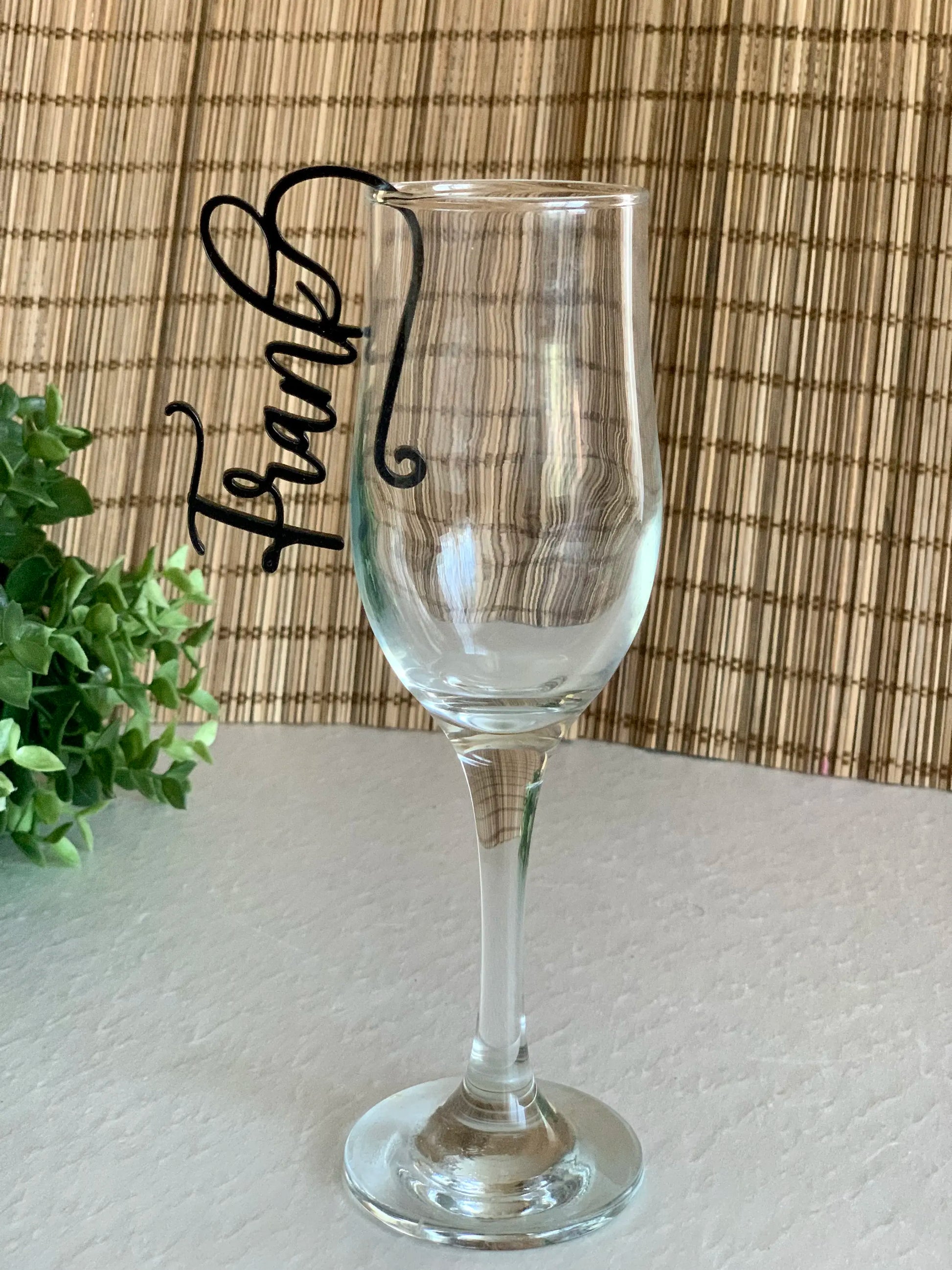 A clear wine glass with a black cursive name tag on the rim, set against a textured bamboo background, with green foliage on the left.