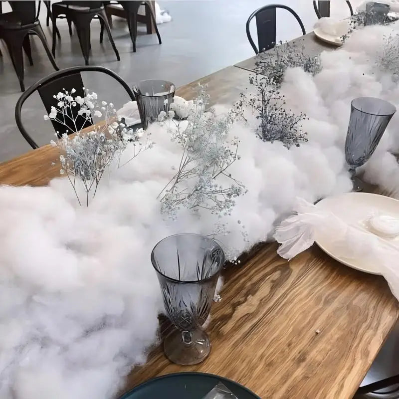 Wooden table set with white cloud-like decor and delicate branches of baby's breath flowers. Grey glass goblets and plates are arranged neatly around the display.