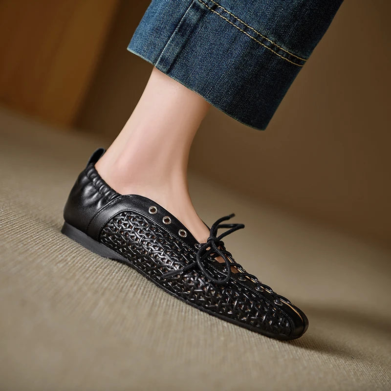 Foot wearing a black woven shoe with metal eyelets and lace-up detail. The background shows beige carpet and blue denim jeans, showcasing modern, casual footwear style.