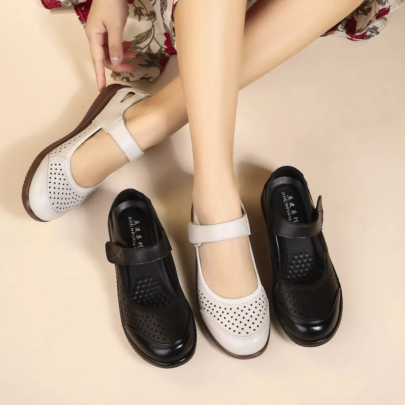 Woman wearing beige perforated Mary Jane shoes, sitting with legs crossed, beside two black similar shoes on beige background. Floral skirt partially visible.